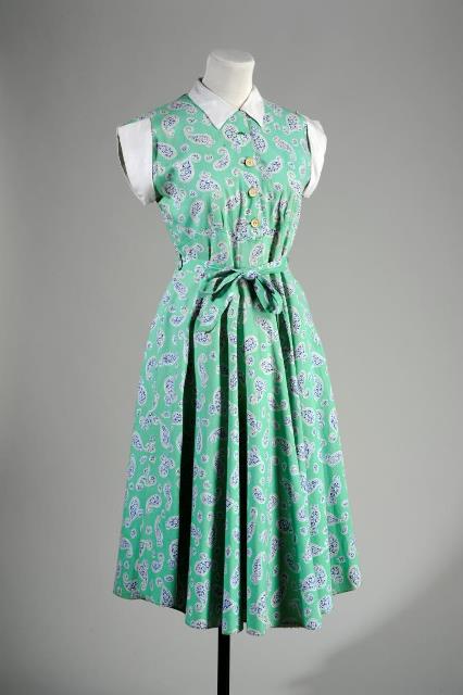 An image of a green ress, mounted on a mannequin. The dress comes below the knee, with a purple pattern, white sleeves and a white collar, with buttons down the middle and a tied bow waist.