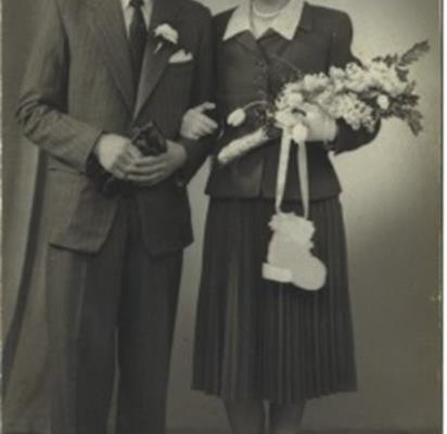 Rosetta and Michael on their wedding day. Rosetta holds a bouquet of flowers, wearing a two-piece suit and skirt with white shoes and pearls. Michael has glasses, and wears a suit and thick rimmed glasses.