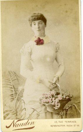 A sepia studio image of a woman dressed in a white dress with a bright red rose on it, holding a basket with flowers in.
