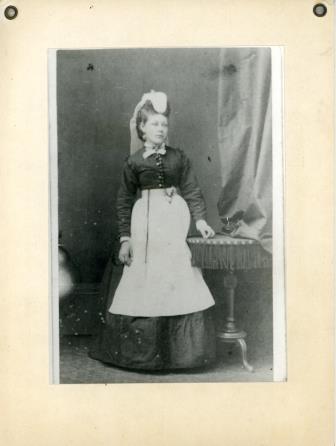 A black and white photograph of a Victorian nurse. She has on a long, grand dress with a white apron, and an extraagant headpiece.