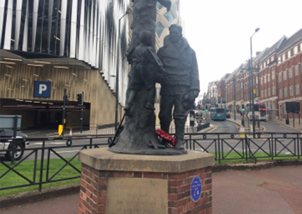 A statue on a brick plinth in the middle of the roundabouts: the statue is of a man iin army gear with a wreath of poppies at his feet.