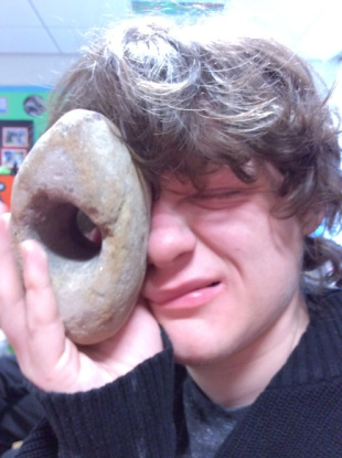 Pupil facing the camera handling a rock with a hole in the middle. He is trying to look at the camera through the hole. He has brown hair and a black sweater.