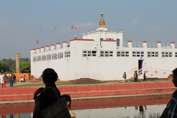 Landscape with a a river and a big white building with red details. There is many windows in the building and a big gold tower on the roof. People are walking around the building and the river.