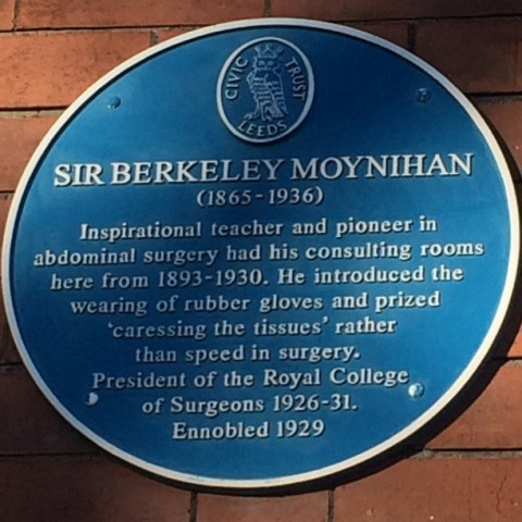 Photo of a red brick wall with a round blue plaque on it. The plaque has the name of Sir Berkeley Moynihan (1865-1936) on it. You can read on the plaque: Inspirational teacher and pioneer in abdominal surgery had his consulting rooms here from 1893-1930. He introduced the wearing of rubber gloves and prized 'caressing the tissues' rather than speed in surgery. President of the Royal College of Surgeons 1926-31. Ennobled 1929.