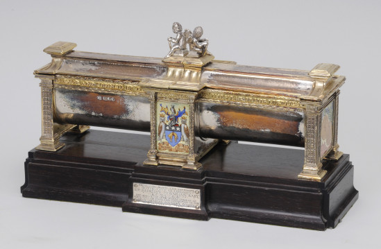 Photo of a beautiful object on a white background. Freedom Casket presented to Sir Berkley Moynihan is gold on a dark wood based. There is the coat of arms of the city of Leeds on the front and writing on the wood base. 