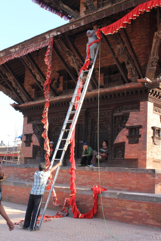 Temple made of red brick with a beautiful detailed dark wood door. 2 people are sat in from of the temple. 2 people are putting decoration on the front of the temple. The decoration are red and gold and they are made of cloth. 1 person is climbing up a long silver ladder to put the decoration up. The other person is holding the base of the ladder.