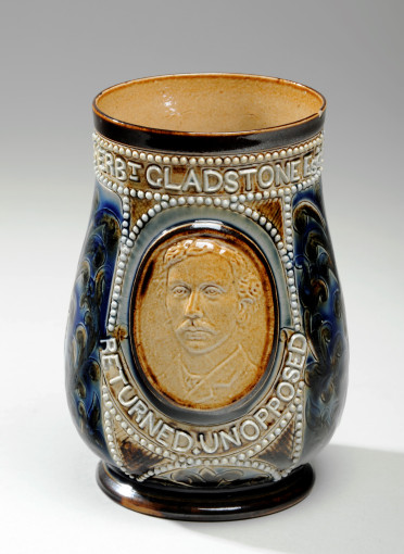 Terracotta Stoneware vase with colourful details. The sides are full of patterns painted with blue, brown, red and white. The vase as the face of a man as the central decoration. Above the man's face you can read Herbert Gladstone and bellow you can read returned unopposed. The written details are white.