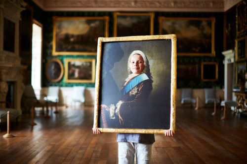 Photograph of a child holding a big portrait in a gold frame with a hole where the face should be. The child has his face in the hole and is posing for the camera. He is standing in the middle of a impressive room at Temple Newsam. Wooden floor, green tapestry and several paintings on the wall. 