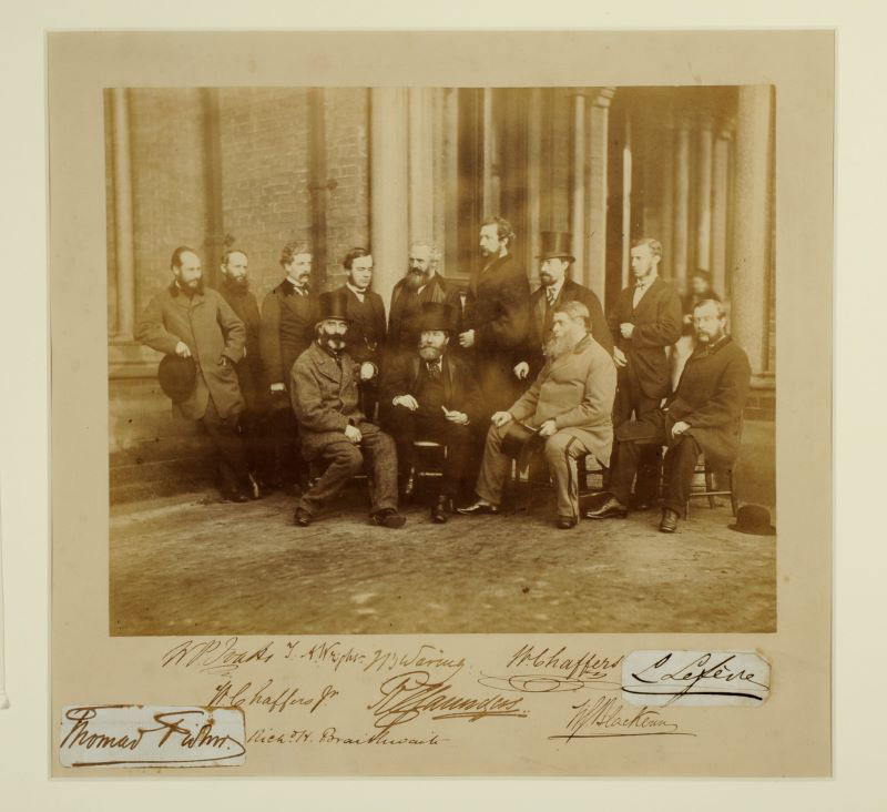 a Black and white photograph of lots of old men sitting on stools to have their photograph taken outside the infirmary. Most of them are wearing hats.