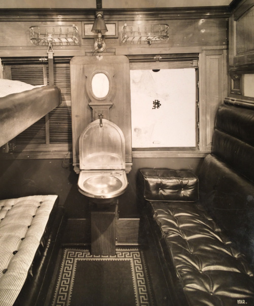A black and white photograph of a sleeping compartment in a train. A sofa is on one side, a bunk bed is on the other and in the middle, what looks like a sink. The compartment is very narrow.