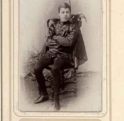 a page of a photograph album featuring a black and white photograph of a young boy dressed all in black with a gnarly cape, sitting with folded arms.