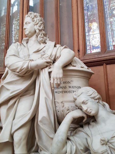 A white, very large statue of a male figure wearing a wig and a woman with her hand resting on her head, looking a bit bored. The man is standing and the woman is sitting, and they are both leaning on a pillar with Wentworth's name inscribed on it.