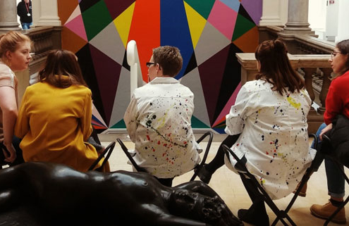 A group of young adults are sat in an art gallery having a chat in front of some modern artwork on the wall by a staircase