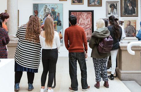 A group of young people are stood in an art gallery looking at painted portraits hung on a wall