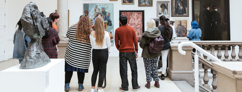 A group of young people are stood in an art gallery looking at painted portraits hung on a wall