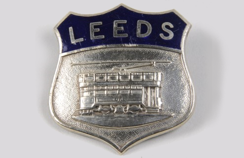 A silver badge with the word 'Leeds' on a blue background and an engraving of a tram