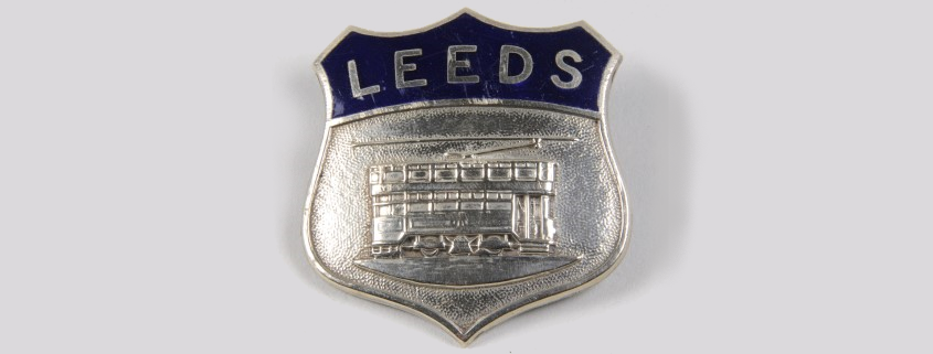A silver badge with the word 'Leeds' on a blue background and an engraving of a tram