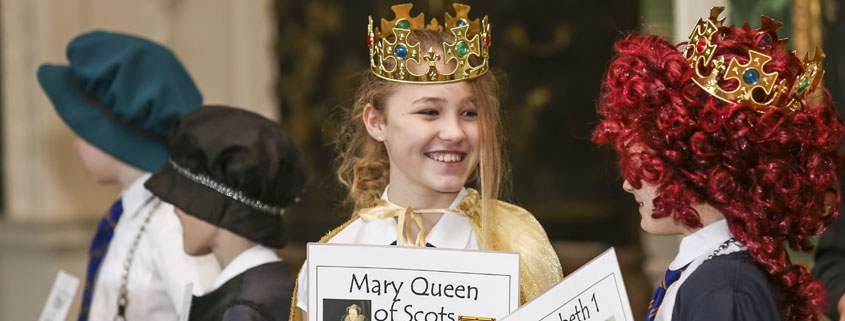 A young girl is dressed in a royal Tudor costume and holding a sheet which says Mary Queen of Scots