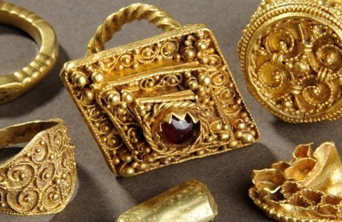 A photograph of 5 ornate gold rings. The middle one is the largest, and has a ruby in the middle.