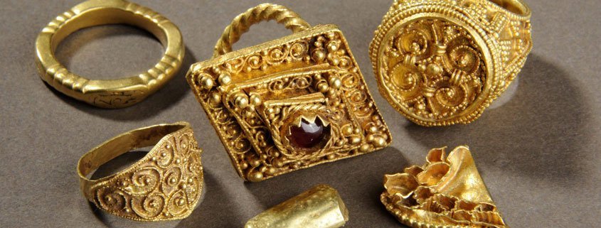 A photograph of 5 ornate gold rings. The middle one is the largest, and has a ruby in the middle.