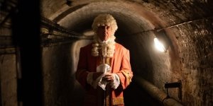 A man is dressed in Georgian clothing, and standing in a dark cellar.
