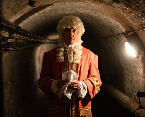 A man is dressed in Georgian clothing, and standing in a dark cellar.