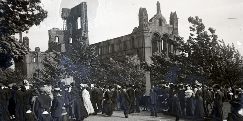 A black and white photograph of a group of Victorian people attending an event, with Kirkstall Abbey in the background