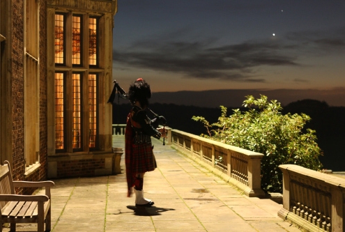 A man dressed in a kilt and a bearskin hat is playing the bagpipes on the terrace at Temple Newsam. It is dark but the sun is beginning to rise.