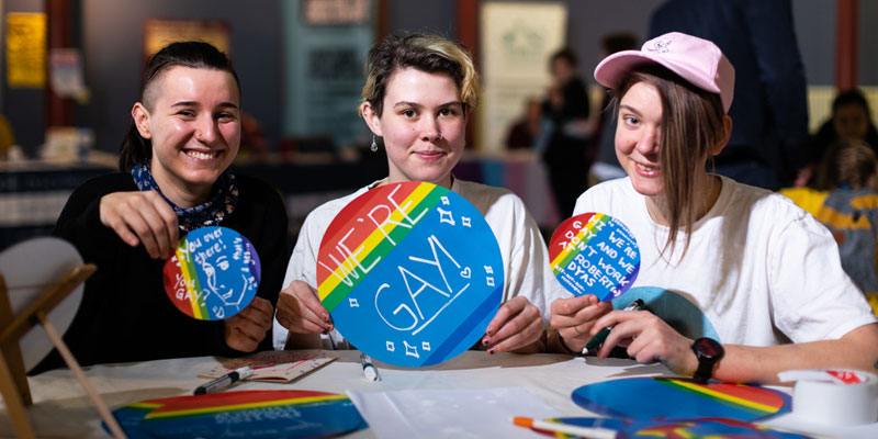 Three people holding up signs saying 'we're gay!' at a previous LGBT+ event at Leeds City Museum