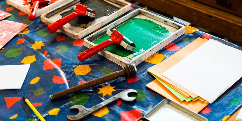 brightly coloured printing materials and tools with cut out shapes laid on a table with a plastic blue tablecloth