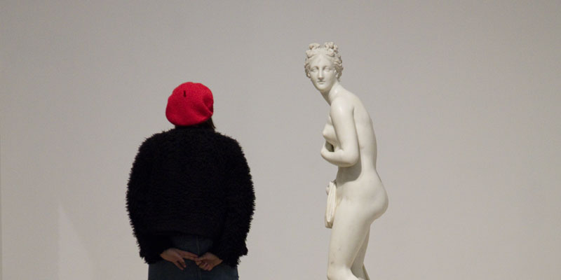 A young woman wearing a red beret and black coat is looking at a classical white sculpture of a naked woman