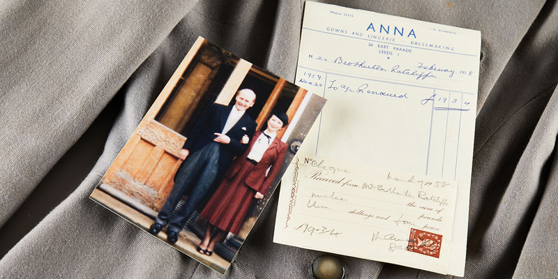 A photograph of two people wearing wedding outfits, with an old fashioned receipt showing the purchase price and item information from ANNA gowns, laid on top of the jacket shown in the photograph
