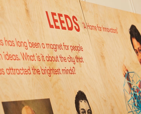 A plywood display board hung on a wall showing details and images for an exhibition on Leeds innovators