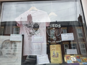A display in someone's lounge window showing t shirts, books, pokemon cards and drawings laid out like a museum window.