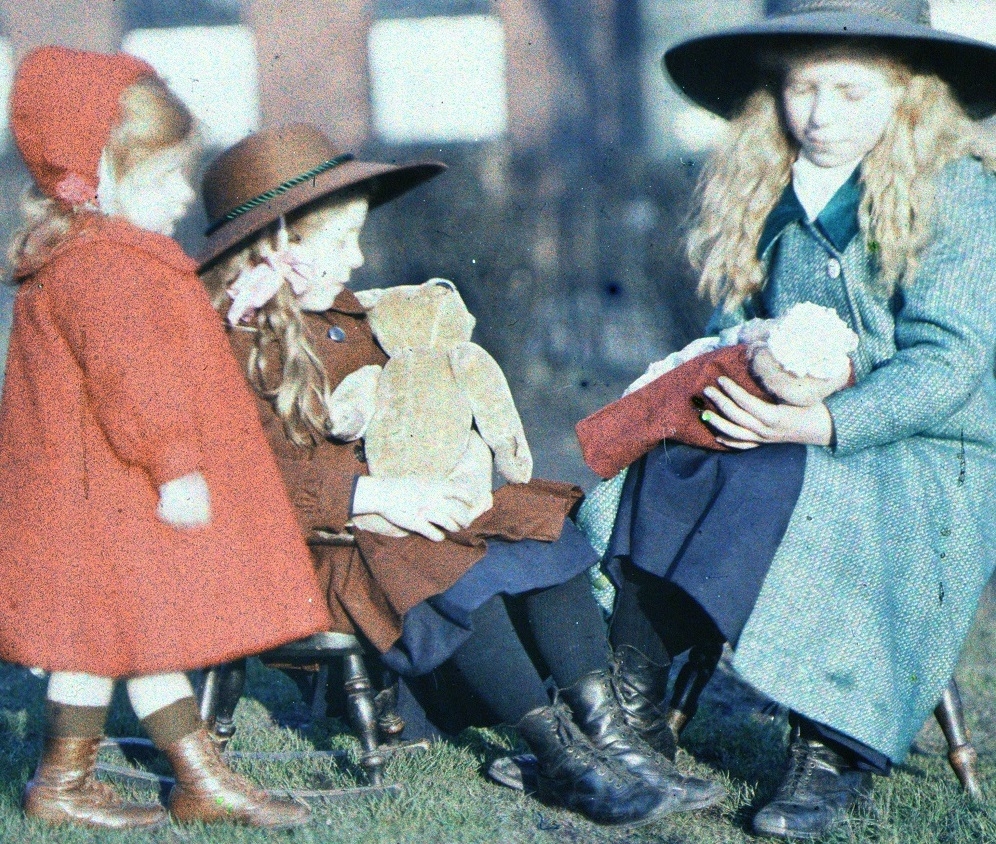 A photograph of 3 young girls. They are all wearing coats and hats and two of them are holding dolls. Two are sat on some stools.