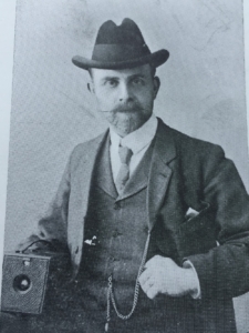 A man in an Edwardian suit is holding a camera and looking at a camera. He looks quite stern and is wearing a hat and a monocle.
