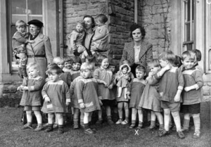 A black and white photograph of children outside a large house with 3 women looking after them,