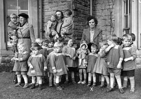 A black and white photograph of children outside a large house with 3 women looking after them,