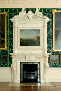 A fireplace and elaborate mantle. Inside the mantle is a painting of a river with a bridge.