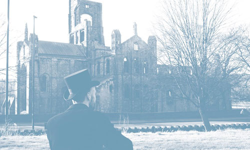A blue coloured photograph of a man in a tophat and dinner jacket sat outside looking over the ruins of an old abbey