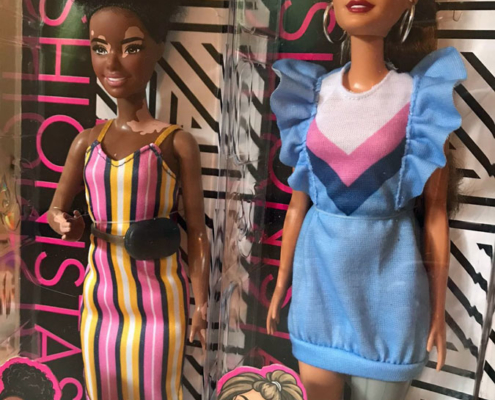 Two Barbie dolls. One has vitiligo, dark curly hair and is wearing a colourful striped dress. The other Barbie doll has a prosthetic leg, wearing a dress with ruffled sleeves and striped pattern