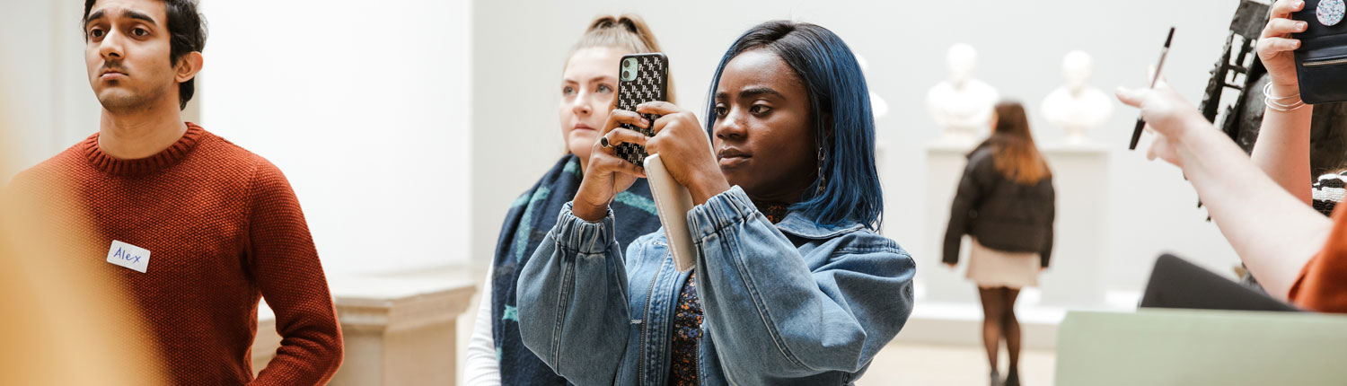 A young girl with blue hair is taking a photograph of something in an art gallery with her phone. There are other young people around her and some sculptural busts in the background
