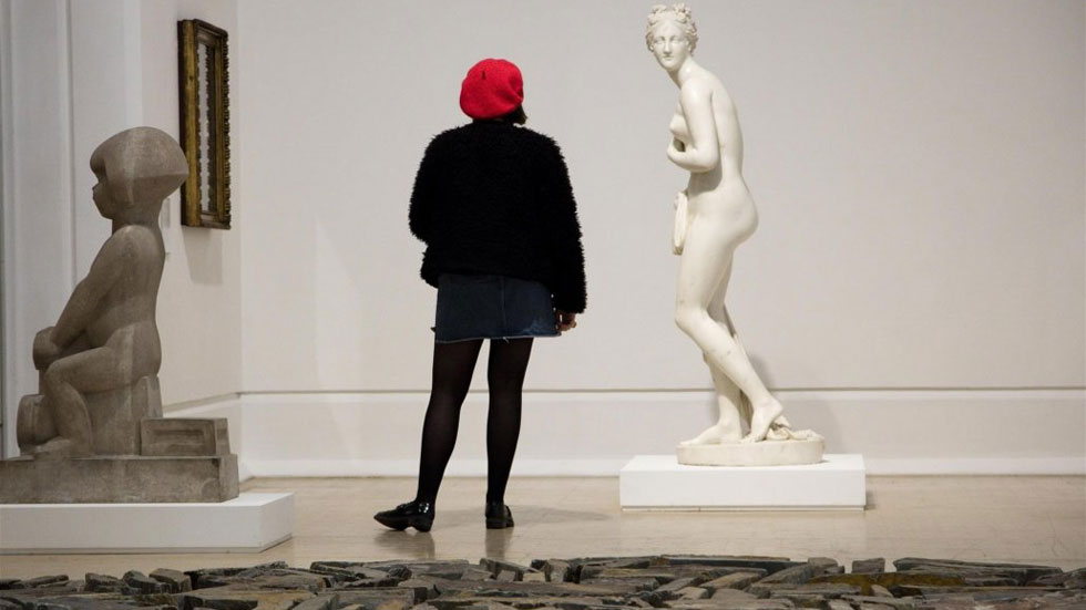 A girl wearing a red beret is looking at sculptures in an art gallery, focusing specifically on a white marble sculpture of a woman covering her naked body