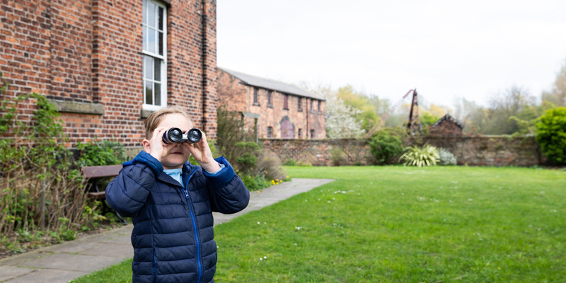 A young boy is stood outside a house looking through some binoculars