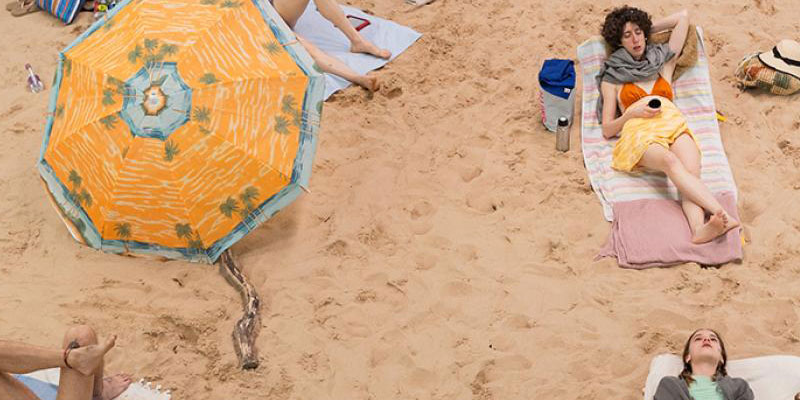A photograph or people sunbathing on a beach with a particular focus on a woman and a large yellow sun umbrella