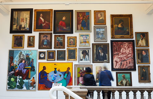 2 visitors are looking at a portrait wall in an art gallery. The works on display range from classical to contemporary paintings, many of which are bright in colour.