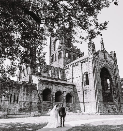 A black and white photo of a wedding in Kirkstall Abbey with the abbey ruins in view