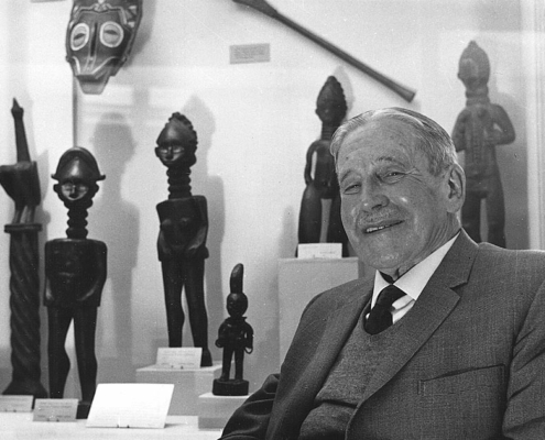 Black and white photo of a white man smiling next to a museum display of African figurines.