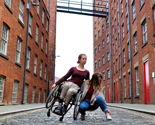 Two people, one in a wheelchair, one crouched down, on a street.