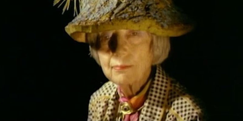 A film still of artist Eileen Agar wearing a decorative hat against a black background